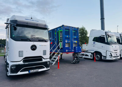 Zwei weiße Elektro-Lkw stehen neben einem mobilen Energiecontainer von FleetBoost auf einem abgesperrten Gelände, eingesetzt zur temporären Stromversorgung und Ladeinfrastruktur.