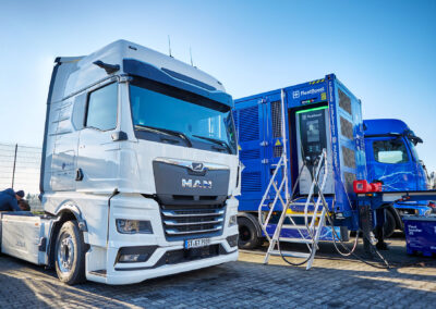 Weißer Elektro-Lkw steht neben einem mobilen Energiecontainer von FleetBoost mit integrierter Ladeeinheit auf einem Betriebsgelände, vorbereitet für die temporäre Stromversorgung und das Laden von Elektrofahrzeugen.