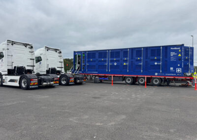 Langer mobiler Fleetboost 40 Batteriecontainer auf Auflieger zwischen zwei weißen Elektro-Lkw auf einem Betriebshof, Ladeeinheit mit Zugangstreppe sichtbar.