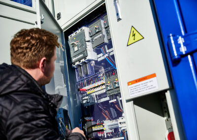 Person überprüft einen geöffneten Schaltschrank eines mobilen Energiecontainers von FleetBoost, sichtbar sind elektrische Komponenten und Sicherheitshinweise zur Hochspannung.
