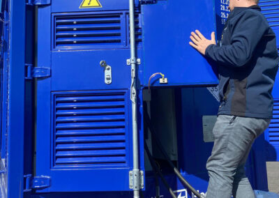 Techniker öffnet die Seitenklappe eines blauen FleetBoost Batteriecontainers auf einem Lkw-Anhänger, sichtbar sind Hochvolt-Warnhinweise und angeschlossene Stromkabel.