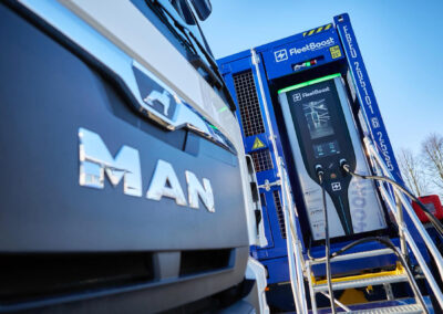 MAN Lkw steht vor einem mobilen FleetBoost Schnellladesystem, das in einen blauen Container integriert ist, mit sichtbarem Display und angeschlossenen Ladekabeln.